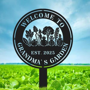 Könnte beinhalten: Schwarzes Metall-Gartenschild mit dem Text "WELCOME TO GRANDMA'S GARDEN EST. 2025" in einem kreisförmigen Design. Das Schild zeigt Silhouetten von Gemüse und ist auf einem Pfahl montiert, vor einem grünen Feld und blauem Himmel.