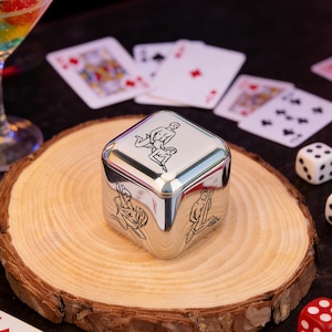 May include: A silver metal dice with black line art illustrations of figures on each side. The dice sits on a wooden slice with playing cards and dice in the background. A cocktail glass is visible in the upper left corner.
