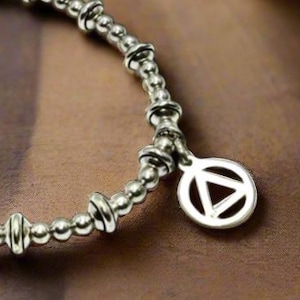 May include: Silver-toned bracelet featuring a series of small, round beads and cylindrical accents. A circular charm with a triangle design hangs from the bracelet. The jewelry is displayed on a wooden surface, with a white flower in the background.