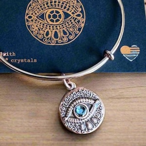 May include: A rose gold-toned Alex and Ani bracelet with an Evil Eye charm. The charm features a blue crystal center and intricate detailing. The bracelet rests on a navy blue card with the brand name and logo, and the words "Adorned with Swarovski crystals".