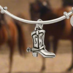 May include: Silver-toned bangle bracelet with a cowboy boot charm. The charm is detailed with a spur and decorative elements. Additional charms include two round pendants with text and symbols. The background features horses and riders.