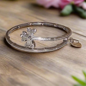 May include: Silver-toned bracelet featuring a decorative cross design. The bracelet has a hinged opening and a small, round charm. The jewelry is displayed on a wooden surface with blurred pink flowers in the background.