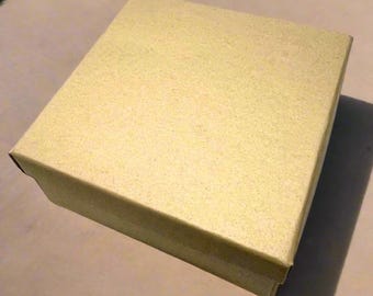 May include: A square, gold-colored cardboard box with a lid. The box has a textured, matte finish and is positioned at an angle, casting a distinct shadow on the surface below. The design is simple, featuring clean edges and a uniform metallic tone.