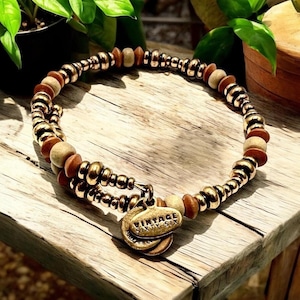 May include: A beaded bracelet with a mix of gold-colored and wooden beads. The bracelet features a double strand design and a small, round charm with the text "VINTAGE SIXTY-SIX". The bracelet is displayed on a wooden surface.