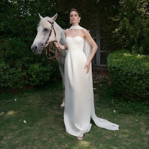 May include: A woman in a white strapless gown with a flowing cape stands next to a white horse. The dress is floor-length and has a fitted bodice. The horse has a brown bridle and reins. The scene is set outdoors with green foliage in the background.
