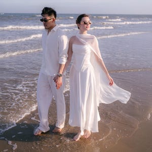 Puede incluir: Una pareja en la playa, ambos vestidos de blanco. La persona de la izquierda lleva una camisa blanca y pantalones, la de la derecha un vestido blanco con una capa fluida. Ambos llevan gafas de sol.