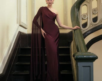 May include: A burgundy one-shoulder gown with a flowing cape detail. The dress is floor-length and made of a silky material. The model is standing on a dark staircase with a green and white banister.