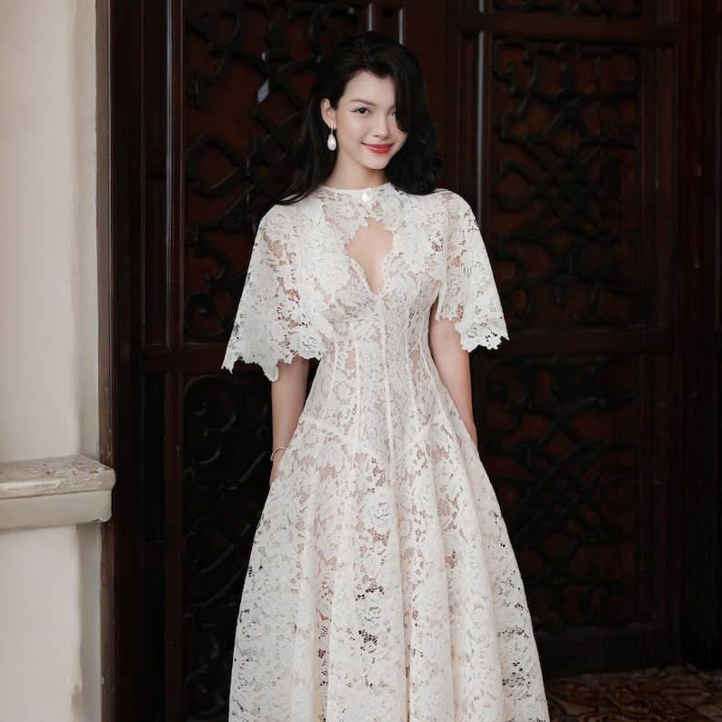 May include: A white lace midi dress featuring a high neckline with a teardrop-shaped cutout, short flared sleeves, and a fitted bodice that transitions into a full, flowing skirt. The intricate floral lace pattern overlays a solid white lining.