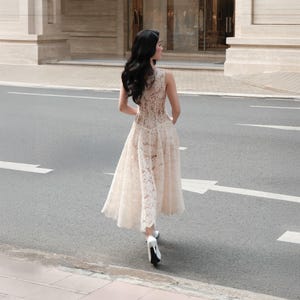 May include: A sleeveless midi dress crafted from cream-colored floral lace featuring a fitted bodice and a flared skirt. The garment is paired with white high-heeled shoes. The dress has a round neckline and a sheer lace overlay throughout the design.