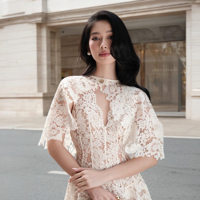 May include: A cream-colored lace dress featuring a high neckline with a central cutout, short cape-style sleeves, and intricate floral patterns throughout the fabric. The garment is constructed from delicate, semi-sheer lace material.