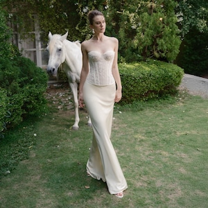 May include: A strapless ivory corset top with delicate lace detailing paired with a matching floor-length satin skirt. A white horse stands in the background, with lush greenery surrounding the scene. The overall aesthetic is elegant and romantic.