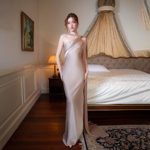 Op de afbeelding: Een beige, vloerlange jurk met een gedrapeerde, asymmetrische halslijn en een hoge split. De jurk heeft een vloeiende sleep. De persoon bevindt zich in een slaapkamer met een bed, een lamp en een schilderij aan de muur.