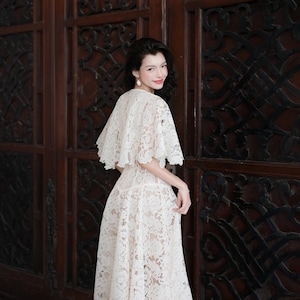 May include: A white lace dress featuring a short-sleeved capelet overlay with a scalloped hemline. The garment is constructed from intricate floral lace fabric that extends from the shoulders down to a flowing skirt with a defined waistline.