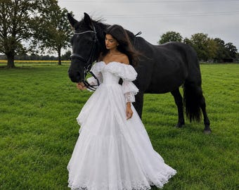 May include: A white off-the-shoulder gown featuring voluminous puff sleeves, a fitted bodice, and a floor-length skirt with tiered layers and intricate lace trim along the hem, standing beside a large black horse in a green grassy field.
