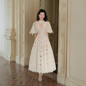 May include: A cream-colored midi-length dress crafted from intricate floral lace. The garment features short, flared sleeves, a high neckline with a central cutout, and a fitted bodice that transitions into a full, flowing skirt with a scalloped hem.