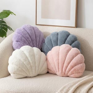 Peut inclure: Quatre coussins en forme de coquillage en peluche, de couleurs lavande, blanc, gris foncé et rose clair. Chaque coussin présente une texture douce et duveteuse avec un design festonné distinct et des segments cousus partant de la base.