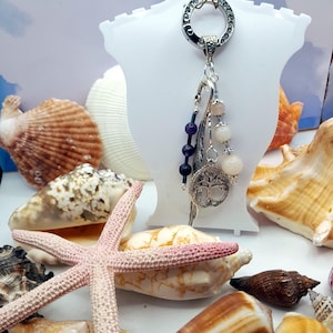 May include: A silver keychain with a decorative ring and dangling charms. The charms include purple and pink beads, a silver feather, and a tree of life pendant. The background features seashells and a pink starfish.