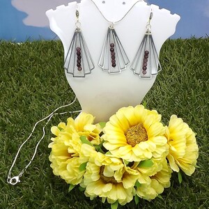 May include: A silver necklace and earring set displayed on a white bust. The earrings and pendant feature a fan-like design with a row of dark red beads. Yellow flowers are in the foreground, with a green grass background.