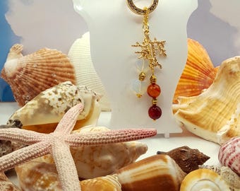 May include: A gold-colored keychain with a tree-like charm and three dangling beads in white, orange, and red. The keychain is displayed on a white stand, surrounded by various seashells and a starfish. The background is a sky-like gradient.