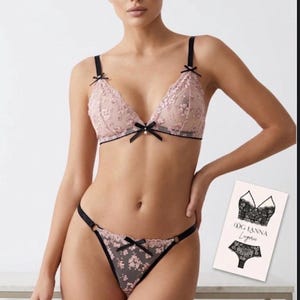 Könnte beinhalten: Ein rosafarbenes Dessous-Set, bestehend aus einem Triangel-BH und einem passenden String. Der BH und der String sind mit floraler Stickerei und schwarzen Schleifen verziert. Das Set wird auf einem Model präsentiert, mit einer kleinen Karte, die das Markenlogo und Produktillustrationen zeigt.