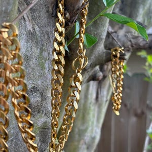 May include: Close-up of gold-colored chain necklaces hanging from a tree branch. The chains have a thick, curb link design. The background includes green leaves and a blurred view of a natural setting.