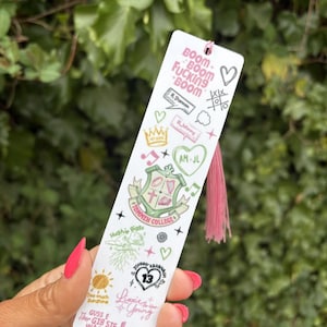 Może przedstawiać: Biała zakładka z różową frędzlą. Zakładka zawiera zielono-biały herb z napisem "Tommen College" oraz inne kolorowe ilustracje, w tym serca, gwiazdy i napis "Boom Boom Fucking Boom".