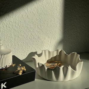 May include: White ceramic bowl with a ruffled edge, featuring a gold ring and a gold charm. The bowl is sitting on a table with a black book and a white candle holder.