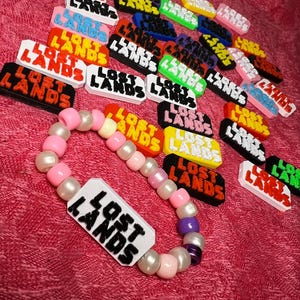 May include: A collection of colorful "Lost Lands" logo charms in various colors, scattered on a pink surface. A beaded bracelet with the "Lost Lands" logo is also present. The charms and bracelet are likely related to music or a festival.