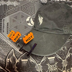 May include: A charcoal gray baseball cap featuring an embroidered white bird logo on the front crown. Two bright orange plastic rectangular attachments with the word HOT printed in black are clipped to the brim with black metal hair clips.