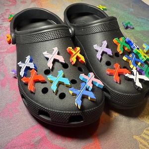 May include: A pair of black clogs adorned with colorful X-shaped charms. The charms are in various colors, including red, orange, blue, pink, and green. The clogs are decorated with multiple charms.