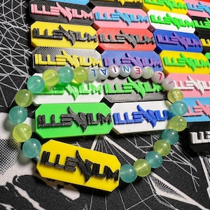 May include: A beaded bracelet with light green and blue beads is displayed in front of a collection of colorful rectangular tags. Each tag has the word "ILLENIUM" printed on it in a contrasting color.