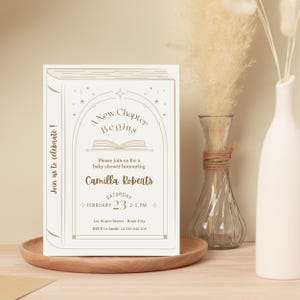 May include: A cream-colored baby shower invitation designed to look like a book. The text reads "A New Chapter Begins" and invites guests to celebrate Camilla Roberts. The date is February 23rd, 2-5 PM.