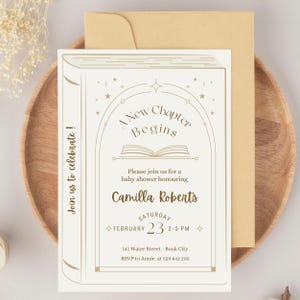 May include: Baby shower invitation card with a book design. The invitation is cream-colored with gold text that reads "A New Chapter Begins" and details for the event. The invitation is on a wooden plate with a gold envelope.