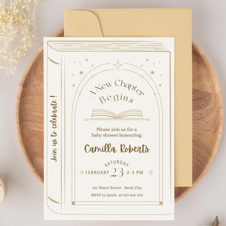 May include: Baby shower invitation card with a book design. The invitation is cream-colored with gold text that reads "A New Chapter Begins" and details for the event. The invitation is on a wooden plate with a gold envelope.