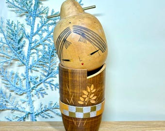Puede incluir: Muñeca de madera artesanal con cabeza en forma de calabaza y base cilíndrica. La cabeza presenta cabello negro pintado y detalles faciales, incluido un pequeño corazón rojo. La base está decorada con un diseño de hojas y una banda a cuadros.