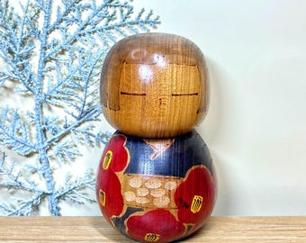 Puede incluir: Muñeca Kokeshi japonesa de madera, con cabeza marrón y cuerpo decorado con flores rojas sobre fondo azul oscuro. La muñeca está sobre una superficie de madera.