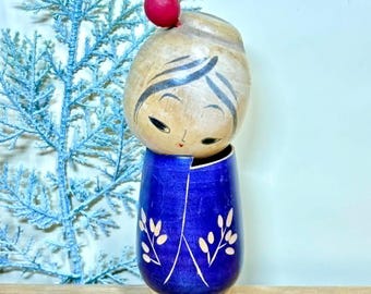 Puede incluir: Muñeca Kokeshi japonesa de madera. La cabeza es de color marrón claro con cabello y rasgos faciales pintados de negro, y una pequeña esfera roja en la parte superior. El cuerpo azul está decorado con diseños florales blancos. La muñeca mide unos 15 cm de alto.