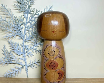 Puede incluir: Muñeca Kokeshi de madera con cabeza redondeada y cuerpo cilíndrico. La cabeza de la muñeca es de color marrón oscuro, mientras que el cuerpo es de un color madera más claro, decorado con diseños florales en tonos naranja y rojo. La muñeca mide unos 15 cm de altura.