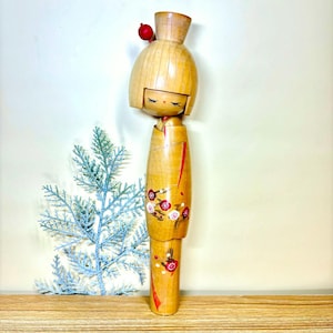 Pode incluir: Boneca Kokeshi de madeira com acabamento castanho claro. A boneca apresenta uma cabeça arredondada com um detalhe vermelho e traços faciais pintados. O corpo é cilíndrico, decorado com linhas verticais vermelhas e desenhos florais. Um item decorativo japonês tradicional.