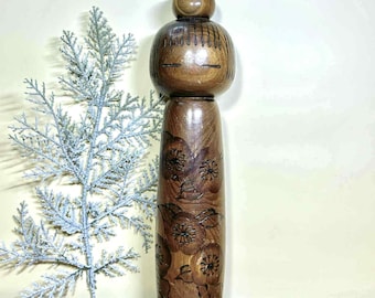 Può includere: Bambola Kokeshi in legno fatta a mano con finitura liscia e lucida. La bambola presenta una testa arrotondata con dettagli del viso incisi e un corpo cilindrico adornato da intagli floreali. La bambola è alta circa 25 cm.