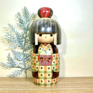 Könnte beinhalten: Eine japanische Kokeshi-Puppe aus Holz mit schwarzer, gefranster Frisur und rotem, gemustertem Kopfschmuck. Der Kimono der Puppe hat ein kariertes Muster in Grün, Orange und Braun mit einer roten Schleife. Die Puppe ist etwa 20 cm groß.