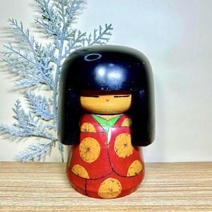 May include: A Japanese Kokeshi doll with a black lacquered head and a red kimono adorned with yellow floral patterns. The doll has a simple face with painted features and a green collar. The doll is displayed on a wooden surface.
