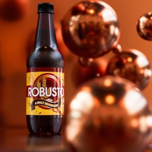 May include: A dark brown bottle of "ROBUSTO" malt beverage with a black cap and a yellow and red label. The label features wheat stalks and a circular design. The background includes blurred bronze-colored spheres.