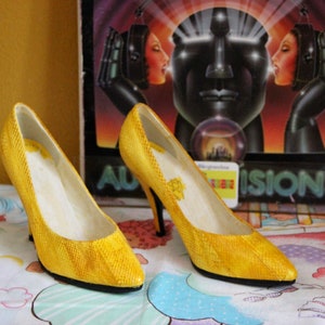 Könnte beinhalten: Ein Paar gelbe High-Heel-Schuhe mit einem strukturierten Stoff. Die Schuhe haben eine spitze Zehenpartie und einen dünnen Absatz.