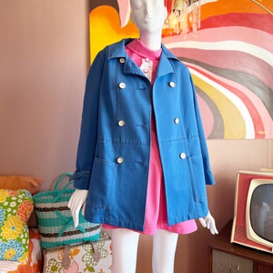 May include: A mannequin displays a vintage blue coat with gold buttons over a pink dress. The coat has large pockets and a classic double-breasted design. The background includes a colorful abstract painting and a retro television.