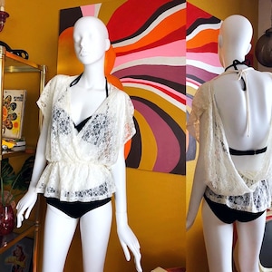 May include: A cream-colored lace top with a wrap-style front and short sleeves. The top is paired with a black bra and black bottoms. The mannequin is displayed in front of a colorful abstract painting.