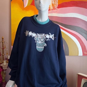 Puede incluir: Sudadera azul marino con cuello verde azulado y un gráfico de un gato en un calcetín, con la palabra "Woody" encima del gato. El gato lleva un suéter verde azulado. La sudadera se exhibe en un maniquí.