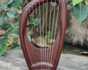 May include: A handcrafted, wooden lyre with a rich, dark brown finish. The instrument features a curved frame, gold-colored strings, and silver tuning pegs. The lyre is displayed against a backdrop of green foliage.