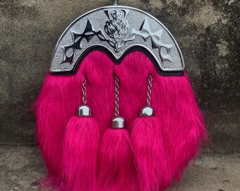 May include: A hot pink sporran with a silver cantle featuring decorative cutouts and a central emblem. The sporran has three matching tassels hanging from the bottom and is made of a fluffy material.