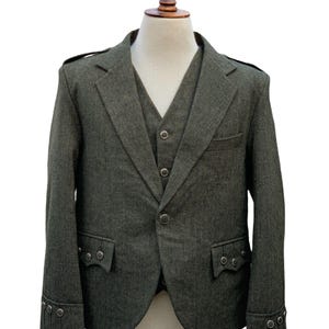 Pode incluir: Um conjunto de casaco e colete de tweed verde, exibido em um manequim. O casaco apresenta lapelas entalhadas, dois bolsos frontais e botões decorativos nos punhos. O colete tem decote em V e fecho de botões. O tecido tem um padrão de espinha de peixe sutil.